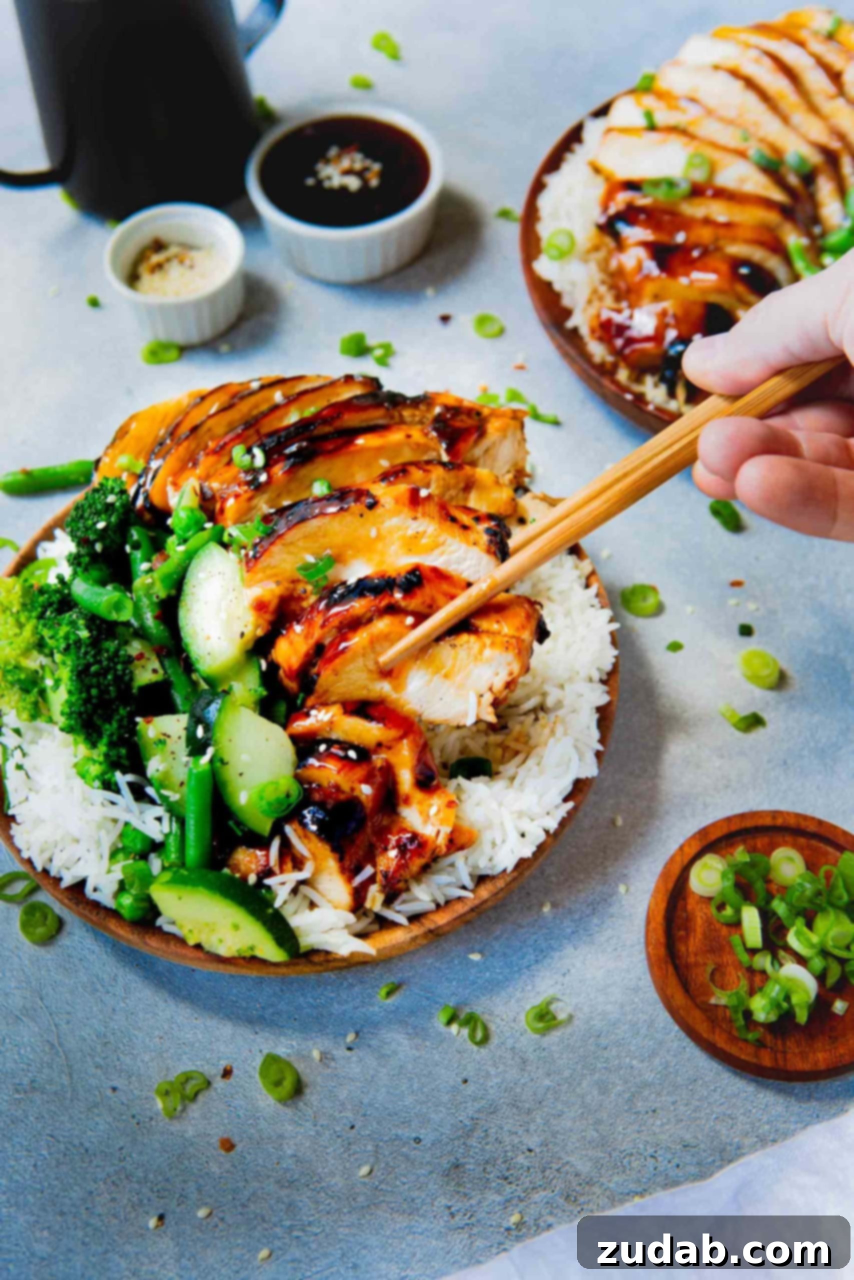 A close-up of a plate of white rice generously topped with saucy teriyaki chicken and bright green steamed vegetables. A pair of wooden chopsticks reaches for a piece of the tender chicken, highlighting its perfect glaze.
