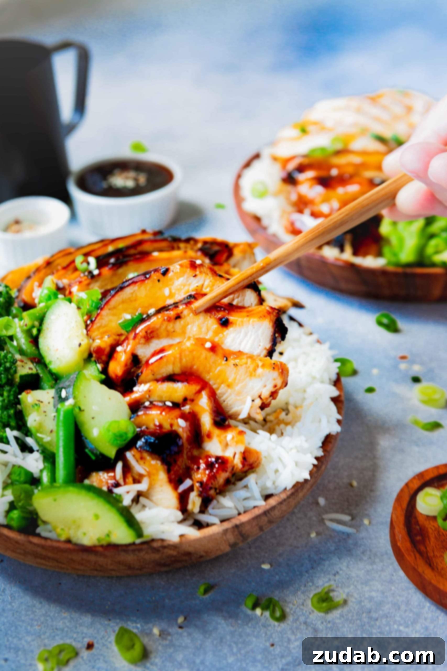 A plate of white rice topped with succulent, saucy teriyaki chicken and vibrant steamed green vegetables. Wooden chopsticks are poised to pick up a piece of chicken, emphasizing its tender texture.