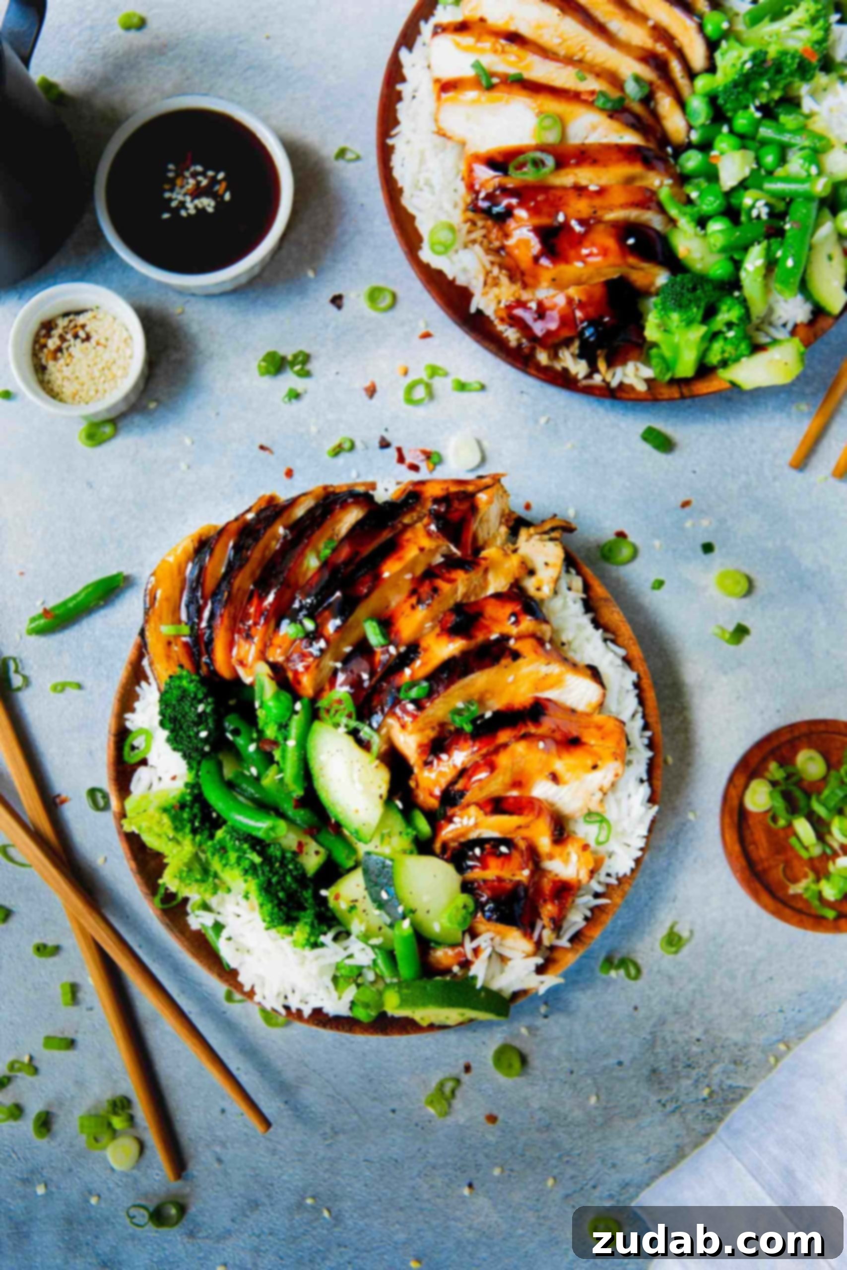 A close-up of a plate of fluffy white rice, adorned with juicy teriyaki chicken, steamed broccoli, zucchini, and green beans. Two wooden chopsticks lie beside the plate, ready for digging in.