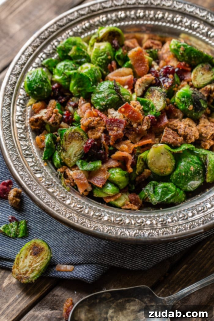 Irresistible Candied Pecan Bacon Brussels Sprouts 3 A vintage, silver bowl sitting on a dark denim napkin with flower full of bright green brussels sprouts that have been halved and pan roasted until golden brown on one side, olive oil and seasoning covering all of them and tossed with red craisins, crisp bacon and crunchy candied pecans and a few pieces of bacon spilled on the table ohsweetbasil.com