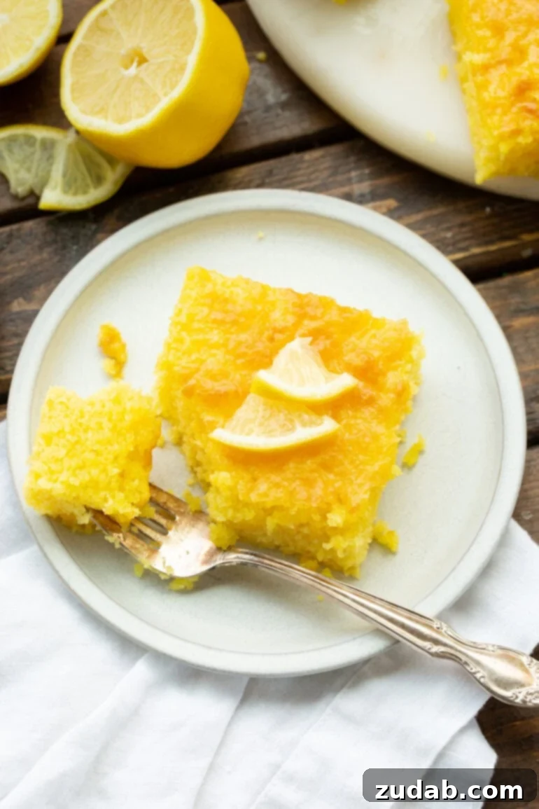 Zesty Lemon Cloud Cake 2 A vibrant slice of moist Lemon Jello Cake on a white dessert plate, with a fork removing a bite, showcasing its tender crumb.