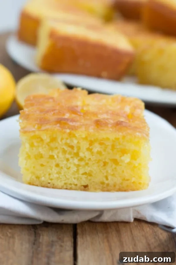 This Lemon Jello Cake is perfect for anytime of the year. The fresh tastes of summer or to brighten the winter holiday meals. A slice of Lemon Jello Cake on a white plate, placed on a white napkin over a wooden table, invitingly presented.