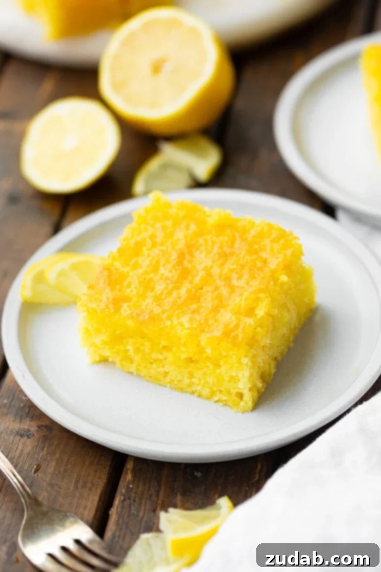 Zesty Lemon Cloud Cake 8 A radiant slice of bright yellow lemon jello cake, perfectly portioned, resting on a small white dessert plate, ready for indulgence.