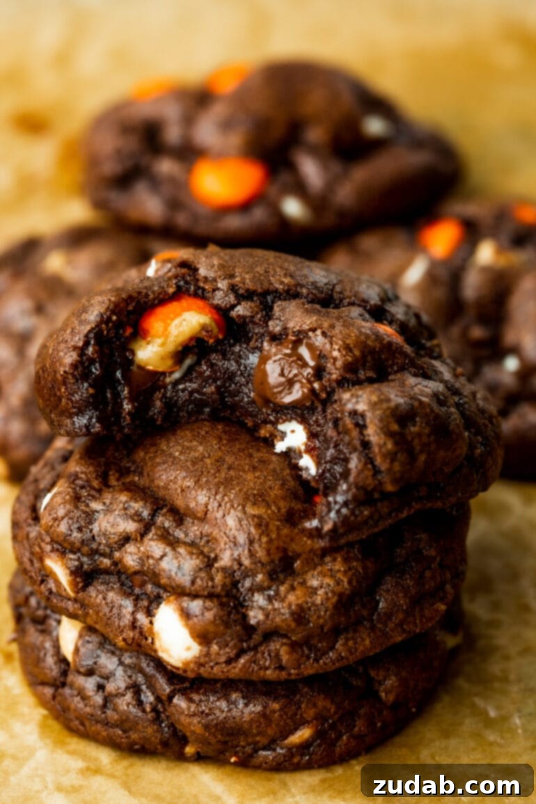 Reese's Triple Chocolate Cookies 2 A photo of three thick, chewy Reese's Triple Chocolate Cookies stacked on top of each other, with a tempting bite taken out of the top cookie, showcasing the melty chocolate chips and Reese's Pieces inside.