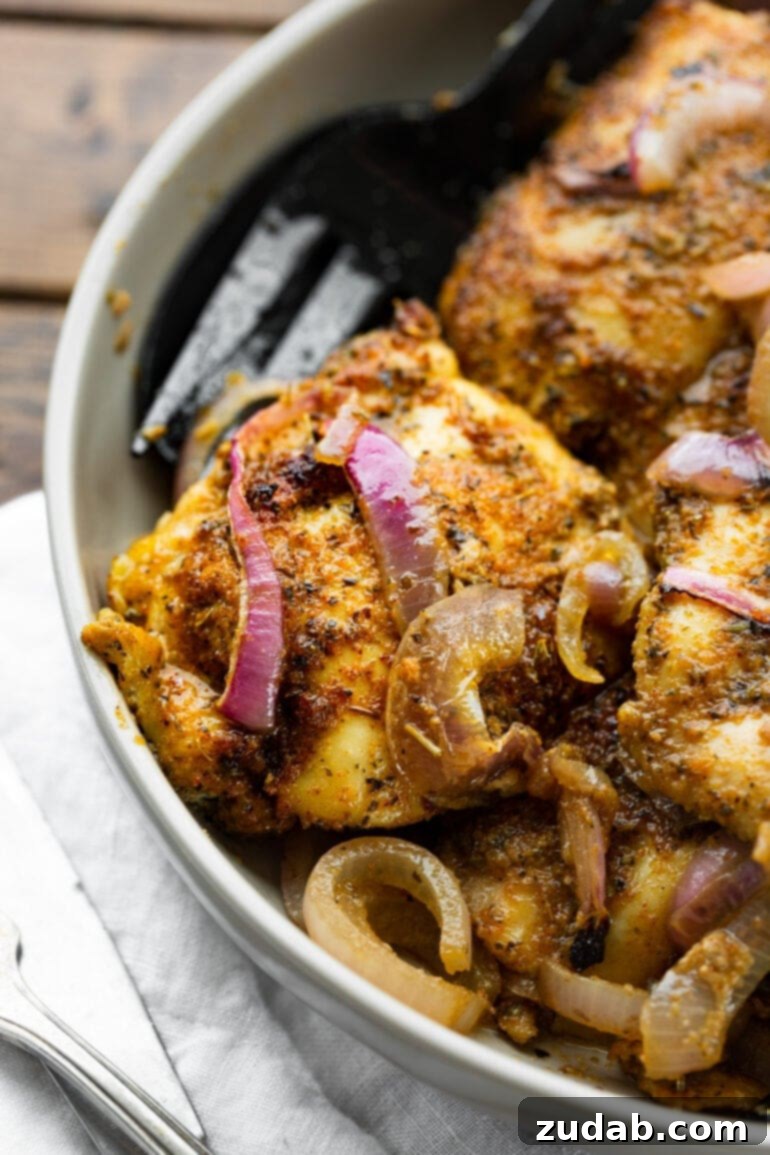 Effortless Baked Boneless Skinless Chicken Thighs 4 A close-up of a perfectly baked chicken thigh, richly seasoned with a dry rub, and adorned with tender slices of red onion.