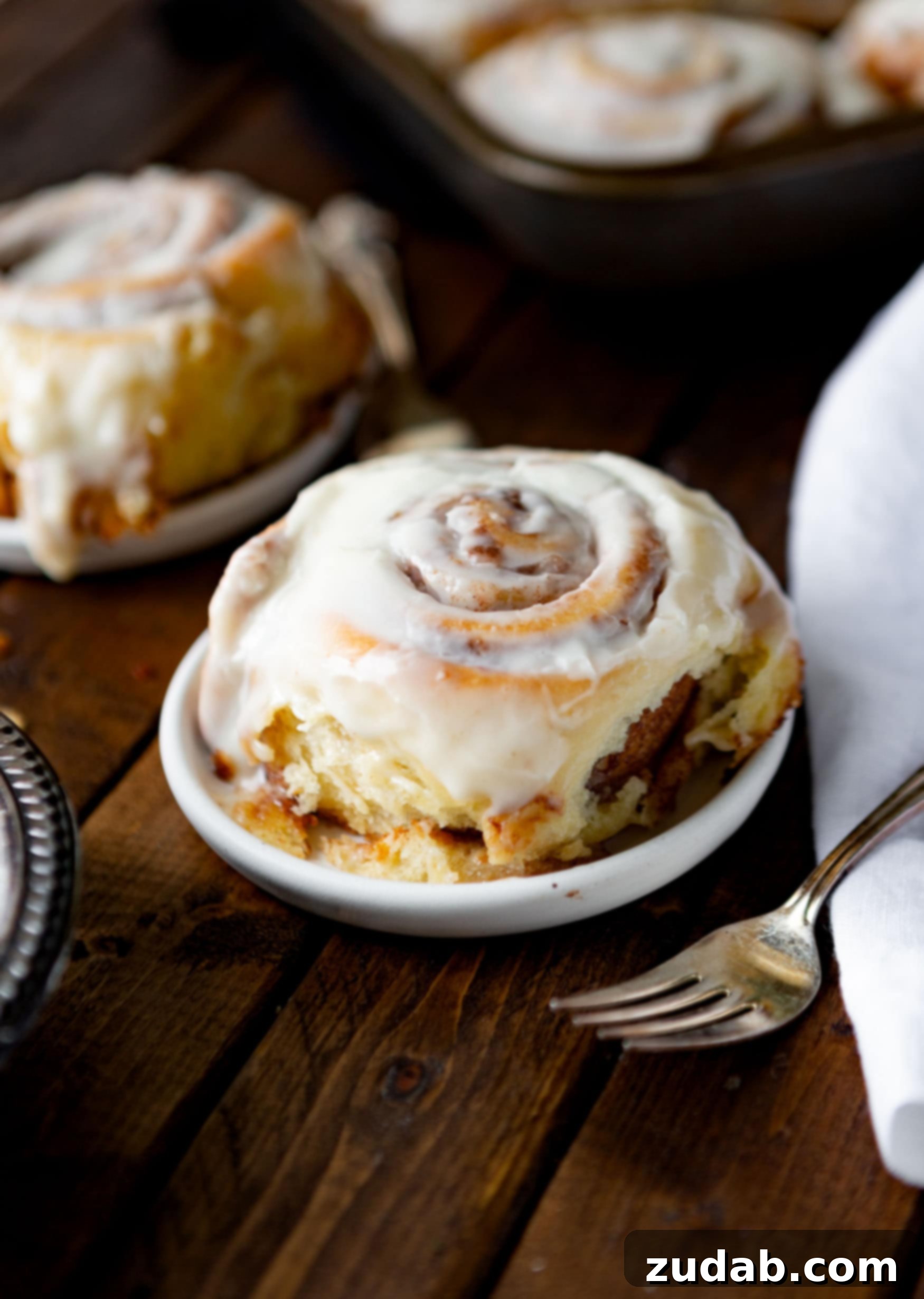 The Definitive Homemade Cinnamon Rolls 2 A photo of a single golden baked cinnamon roll on a small dessert plate, with a silver fork resting beside the plate, emphasizing its readiness to be eaten.