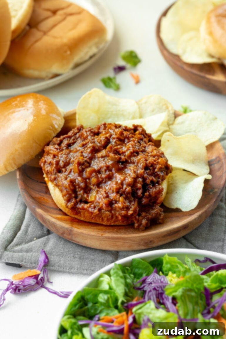 The Ultimate Sloppy Joe Recipe 3 A close-up shot of a sloppy joe sandwich with the top bun removed, showcasing the generous, perfectly seasoned ground beef filling, surrounded by a scatter of crispy potato chips.