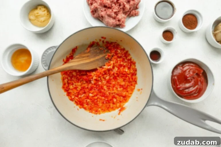 The Ultimate Sloppy Joe Recipe 4 A Dutch oven on the stove, filled with diced onions and red bell peppers being sautéed in butter, with other sloppy joe ingredients like ground beef and spices laid out around the pot.