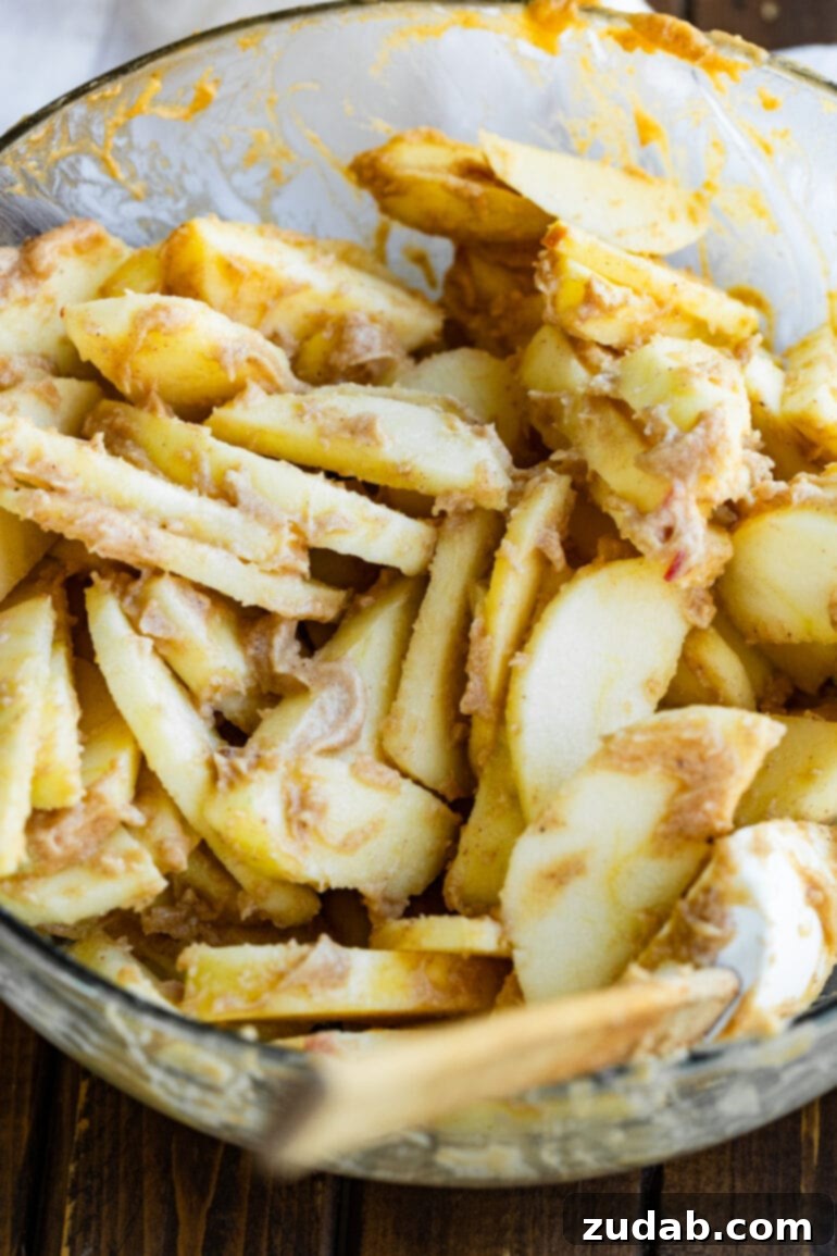 The Ultimate Homemade Apple Crisp 3 A large glass bowl filled with expertly peeled and thinly sliced apple pieces, glistening with a creamy, sugary, and cinnamon-infused coating, ready for baking into the perfect apple crisp.