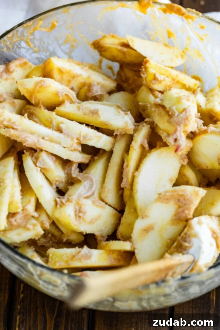 The Ultimate Homemade Apple Crisp 7 A large glass bowl brimming with peeled and thinly sliced apple pieces, beautifully coated in a creamy, cinnamon-infused sugar mixture, perfectly prepped for baking into a delicious apple crisp.