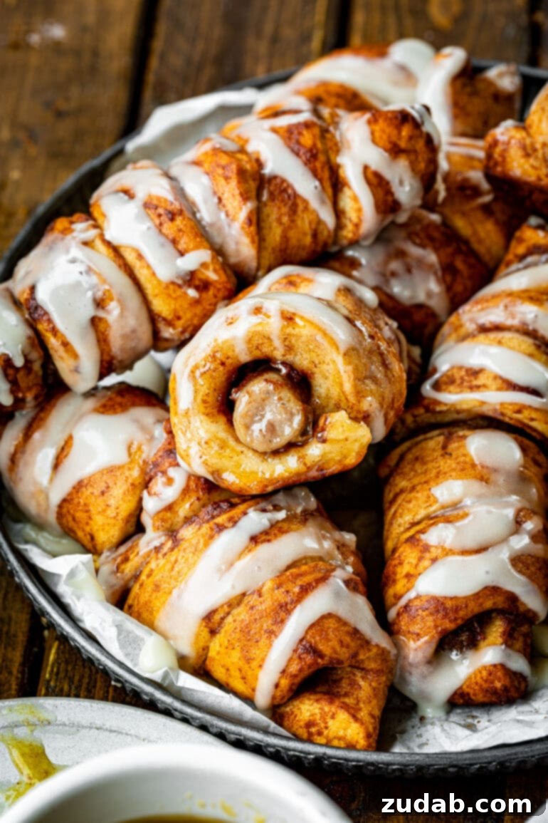 Pigs in a Sweet Blanket 2 A vibrant photo showcasing several golden-brown cinnamon roll pigs in a blanket, each wrapped around a sausage link and topped with a creamy vanilla glaze. They are arranged appealingly, suggesting a delicious breakfast or brunch.