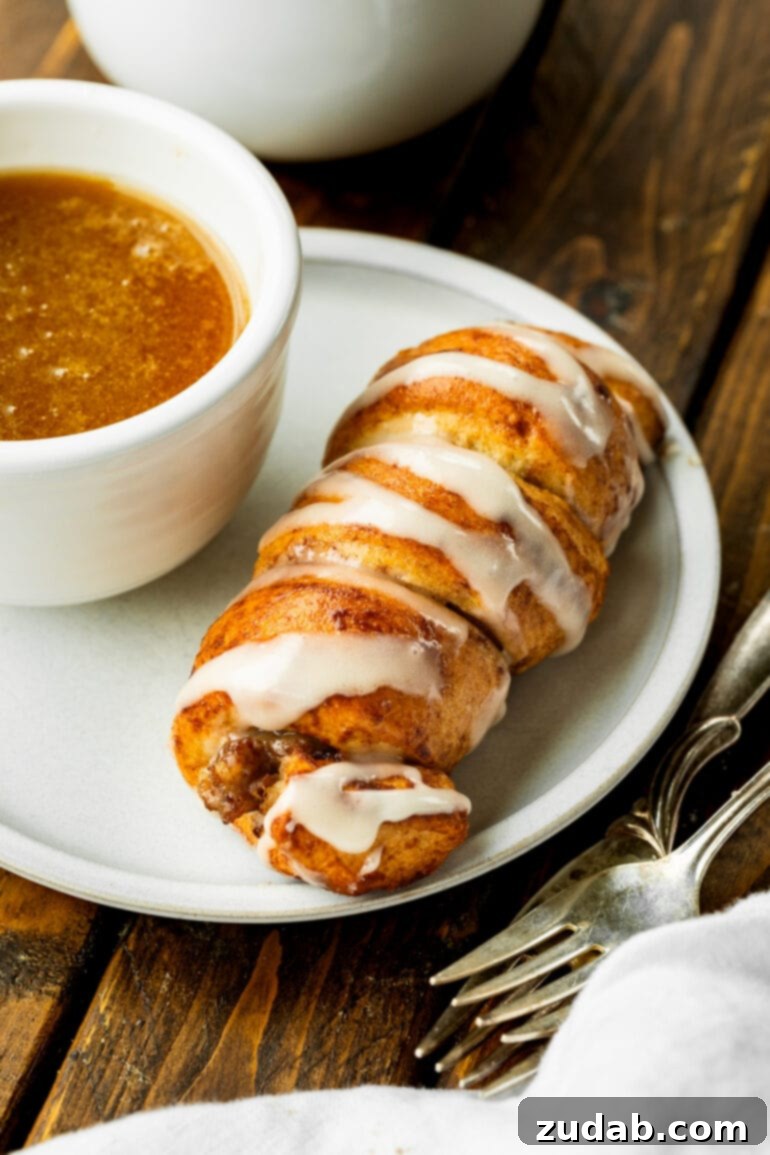 Pigs in a Sweet Blanket 5 A close-up shot of a perfectly baked cinnamon roll pig in a blanket resting on a white plate, positioned next to a small, elegant cup filled with the rich maple butter dipping sauce. This image emphasizes the readiness for dunking.