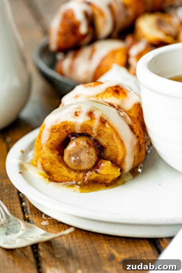 Pigs in a Sweet Blanket 6 A mouth-watering close-up of a single cinnamon roll sausage dunker, golden and perfectly baked, resting next to a small bowl of rich maple butter syrup, inviting a delicious dip.