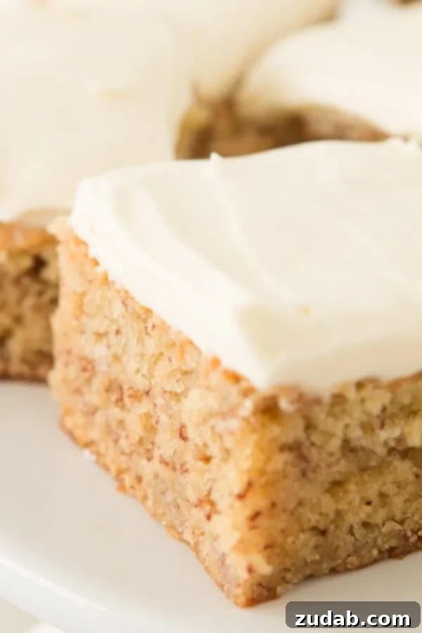 A close-up of a slice of moist banana cake, topped with creamy, smooth cream cheese frosting, highlighting its enticing texture.