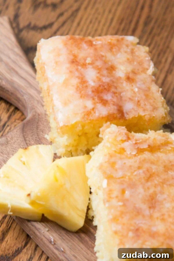 Pineapple crush jello cake is seriously one of the most moist cakes out there. The pineapple niblets are barely noticeable but give such flavor! A rustic wooden cutting board displays several slices of moist Island-Inspired Jello Pineapple Cake, beautifully drizzled with a glossy coconut glaze and artfully surrounded by juicy chunks of fresh pineapple, creating a vibrant tropical presentation.