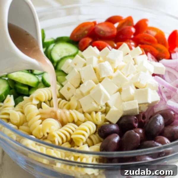 A fresh and easy Greek Pasta Salad just in time for summer! This crowd-pleasing side dish is tasty with grilled meats and at all your backyard barbecues. This Easy Greek Pasta Salad is all about one thing: The chunks of feta cheese. Yes, I love the chewy pasta, the crunch of the fresh veggies, and the tangy dressing. It's basically summer in a bowl, colorful and fresh. Pouring red wine vinaigrette into a bowl full of Greek Pasta Salad ingredients.