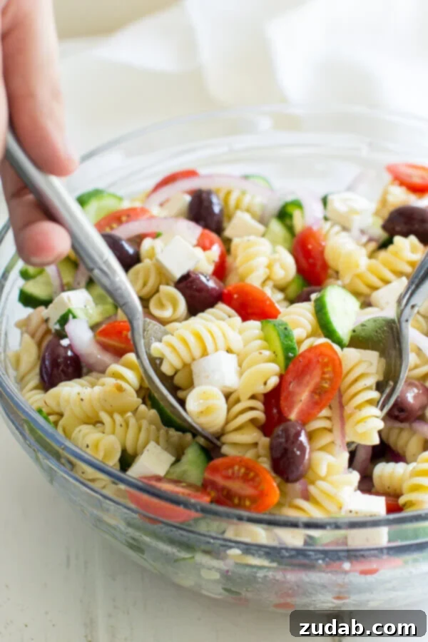 A fresh and easy Greek Pasta Salad just in time for summer! This crowd-pleasing side dish is tasty with grilled meats and at all your backyard barbecues. This Easy Greek Pasta Salad is all about one thing: The chunks of feta cheese. Yes, I love the chewy pasta, the crunch of the fresh veggies, and the tangy dressing. It's basically summer in a bowl, colorful and fresh. Grabbing two scoopfuls of Greek Pasta Salad in a clear bowl.