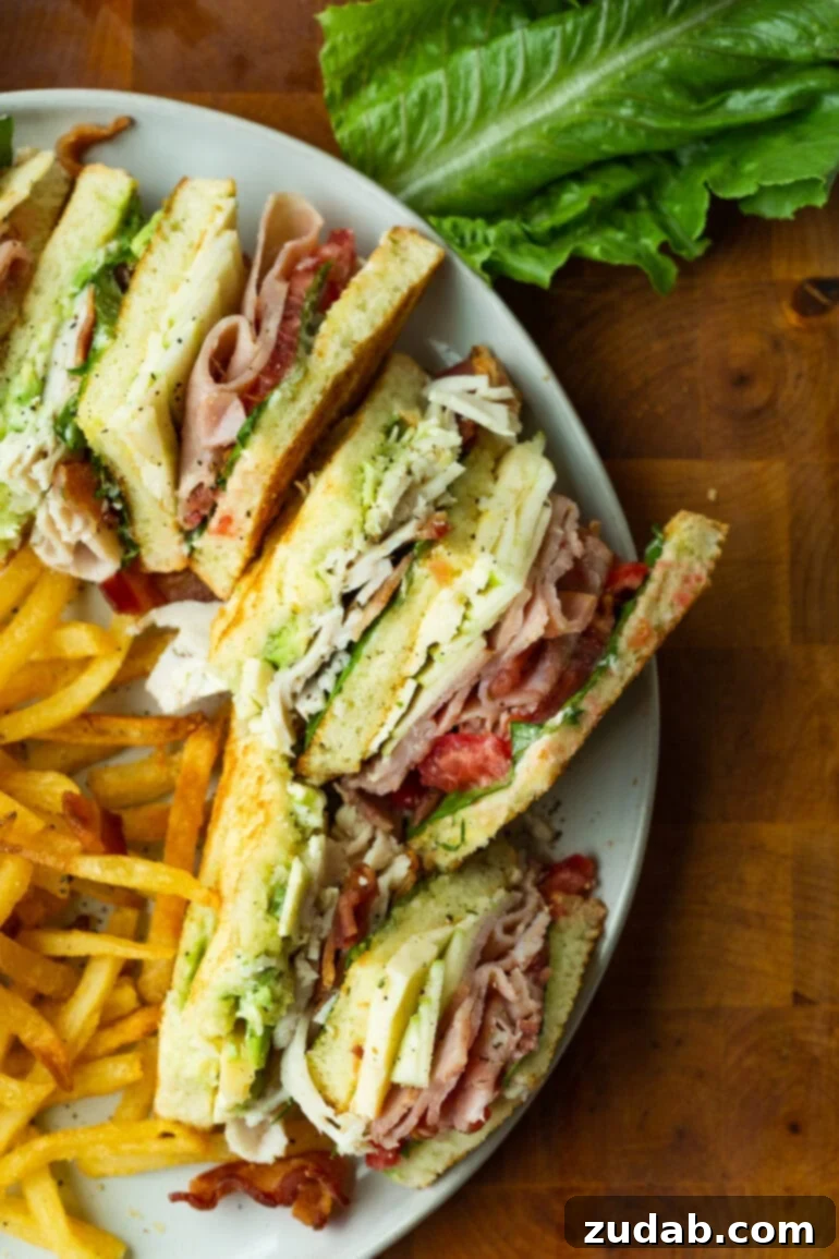 The Ultimate Triple Decker Club Sandwich 9 a photo of several sections of a club sandwich sitting on a white dinner plate with a side of crispy french fries.