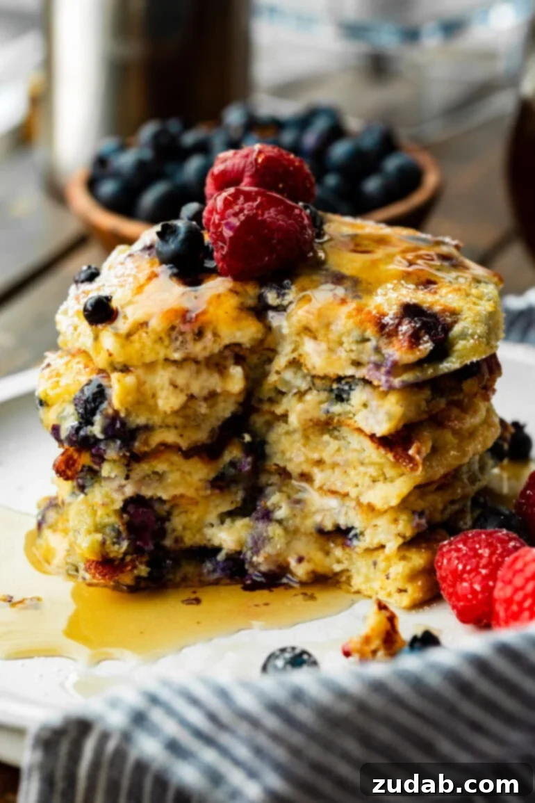 Protein-Packed Wild Blueberry Cottage Cheese Pancakes 2 A photo of a stack of 5 fluffy blueberry pancakes with a bite taken out, topped with fresh berries and covered with syrup, emphasizing their wholesome appeal.