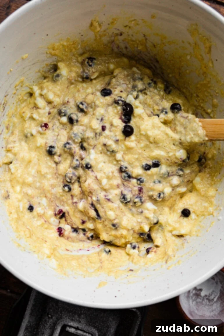 Protein-Packed Wild Blueberry Cottage Cheese Pancakes 5 A photo capturing the vibrant batter for cottage cheese blueberry pancakes being gently mixed in a bowl with a wooden spoon, showcasing the rich texture and inclusion of fresh blueberries.
