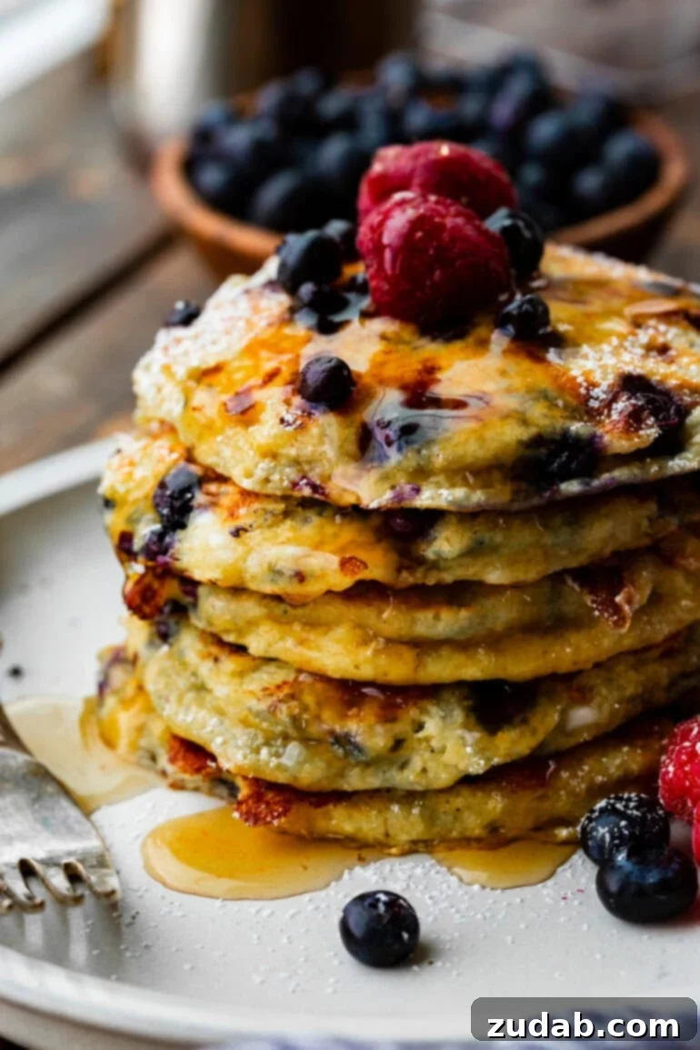 Protein-Packed Wild Blueberry Cottage Cheese Pancakes 9 A photo of a stack of fluffy blueberry cottage cheese pancakes, generously covered in maple syrup and adorned with fresh raspberries and blueberries, ready to be enjoyed.