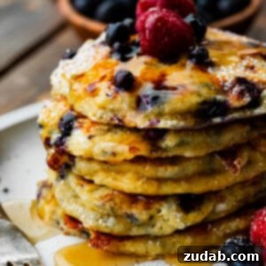 A photo of a stack of fluffy blueberry cottage cheese pancakes, generously covered in maple syrup and topped with fresh raspberries and blueberries.