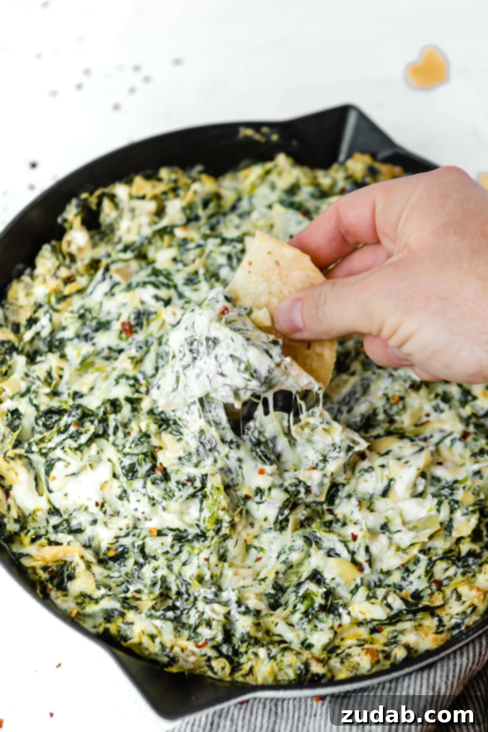 Cheesy Spinach Artichoke Dip A cast iron skillet filled with spinach and artichoke dip. The spinach and artichoke are mixed into a white creamy cheese sauce and someone is dipping a tortilla chip into the dip.