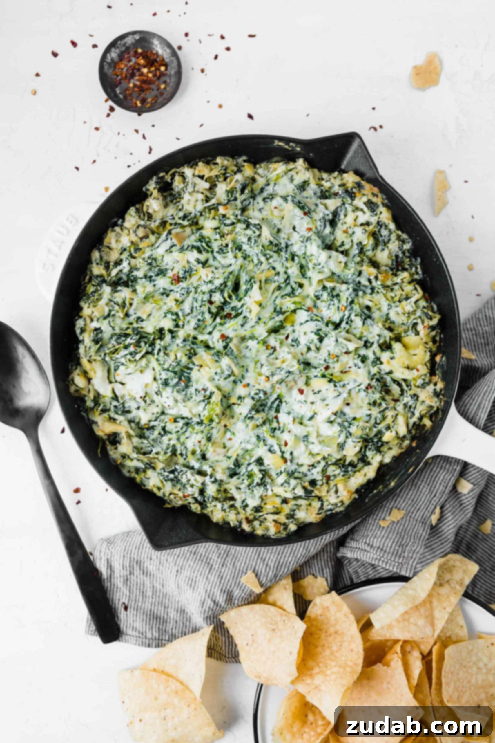 Cheesy Spinach Artichoke Dip A cast iron skillet with spinach and artichoke dip. It is creamy and has green spinach and artichokes mixed with the white cheesy sauce. There are tortilla chips and a spoon next to the dip.
