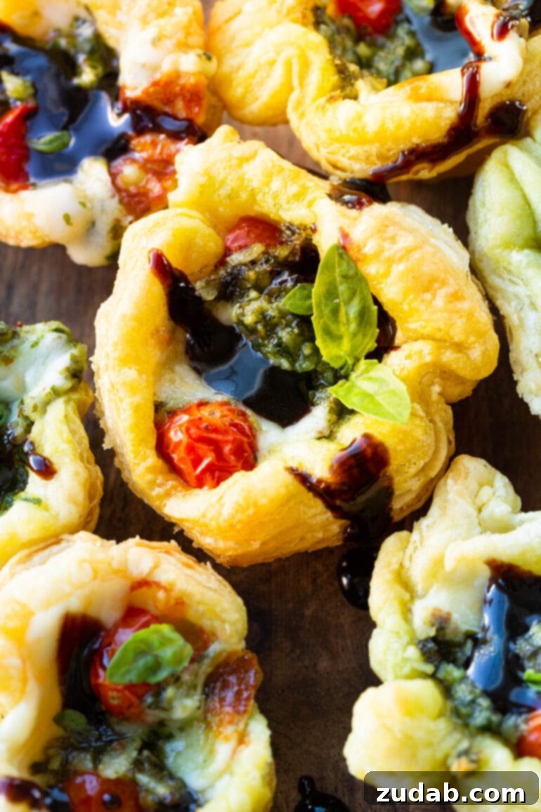Speedy Cheesy Caprese Pesto Tarts 11 A close-up photo of a golden, crispy pesto puff pastry bite, elegantly topped with a glistening balsamic glaze and fresh basil leaf. This image emphasizes the appetizing texture and gourmet presentation of the tarts.