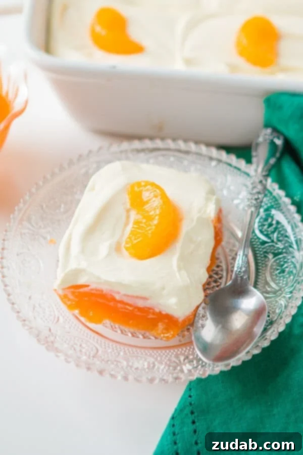 A classic Lemon Mandarin Orange Jello Salad, perfect for holiday meals, featuring a bright orange gelatin base and a creamy lemon topping. slice of jello fruit salad on glass plat with spoon