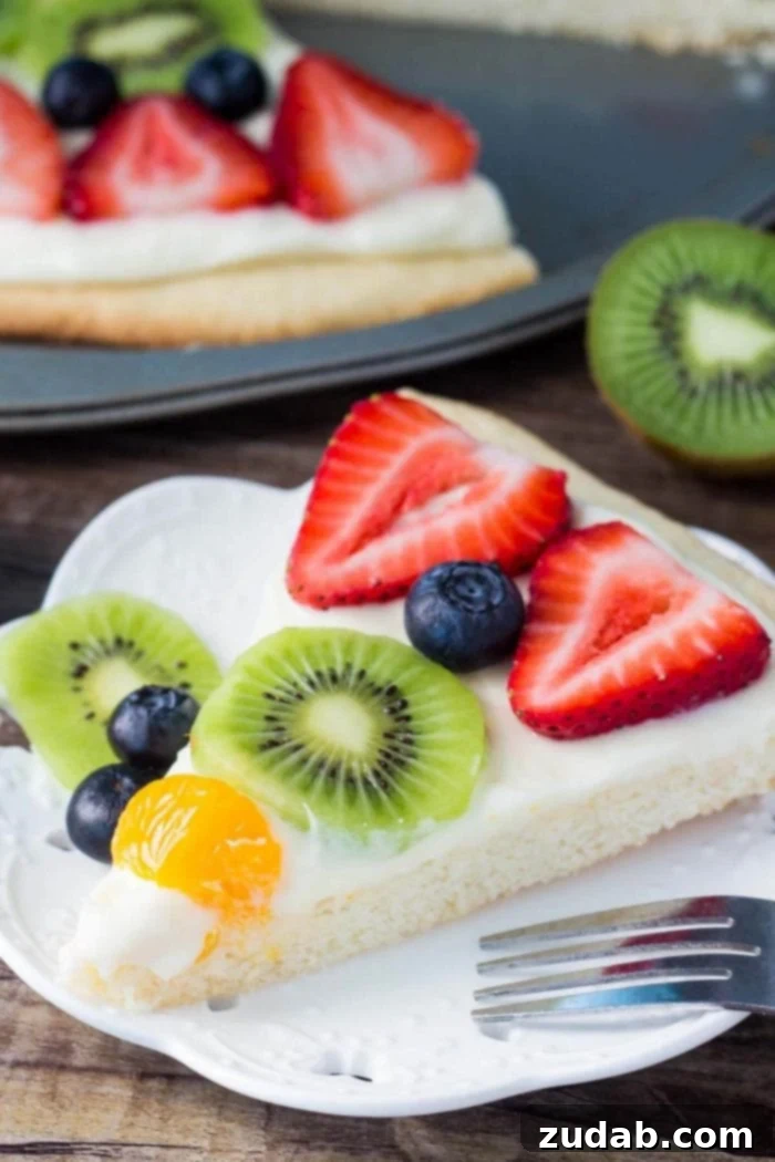 This sugar cookie fruit pizza is perfect for summer. Decorated with tangy cream cheese frosting and fresh fruit - it looks and tastes absolutely delicious. slice of sugar cookie fruit pizza on white plate with fork, showing layers of cookie, frosting, and fruit