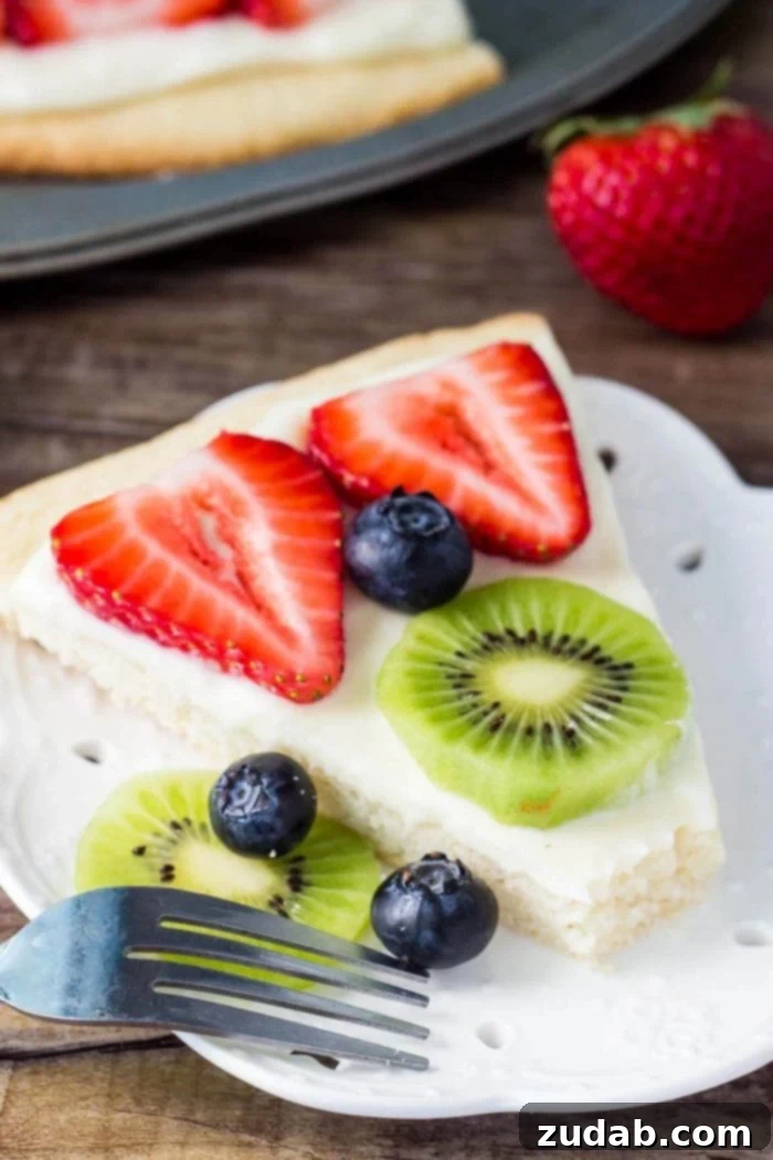 This sugar cookie fruit pizza is perfect for summer. Decorated with tangy cream cheese frosting and fresh fruit - it looks and tastes absolutely delicious. slice of sugar cookie pizza topped with fruit on white plate, ready to be enjoyed