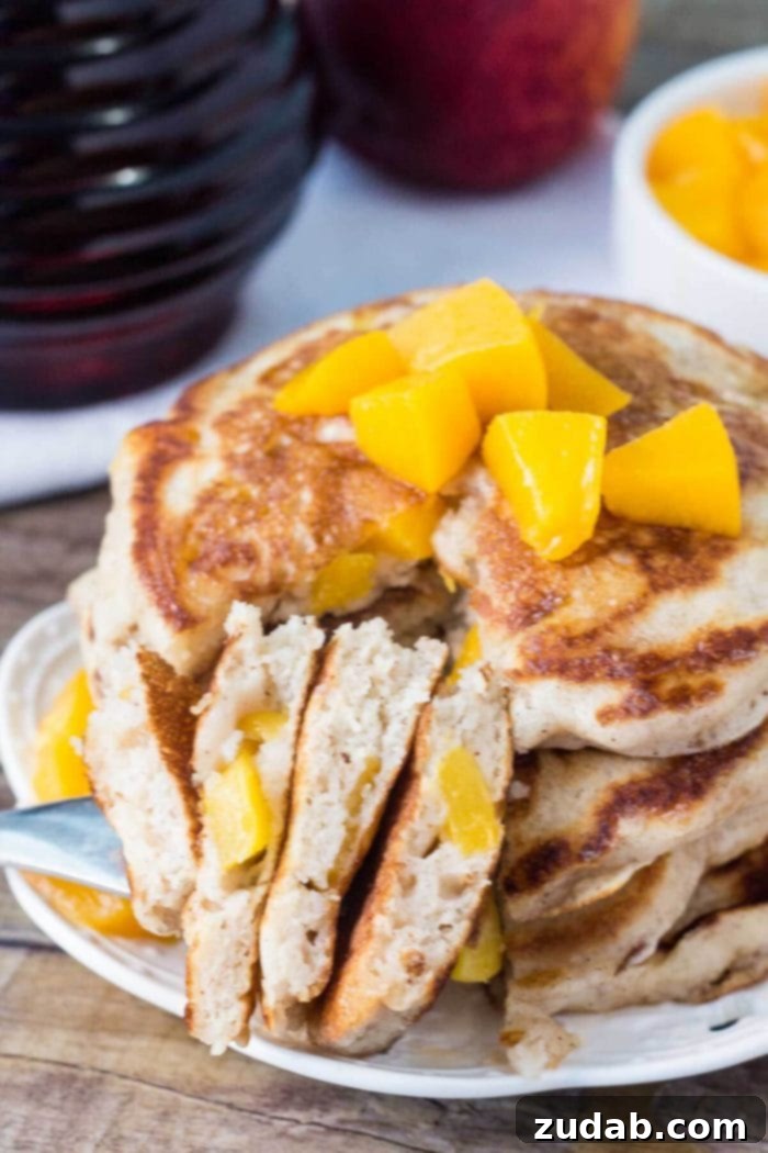 Cut peach pancakes, showcasing their delicious interior. A stacked tower of fluffy peach pancakes, cut into halves, revealing juicy peach pieces inside and a sprinkle of powdered sugar.