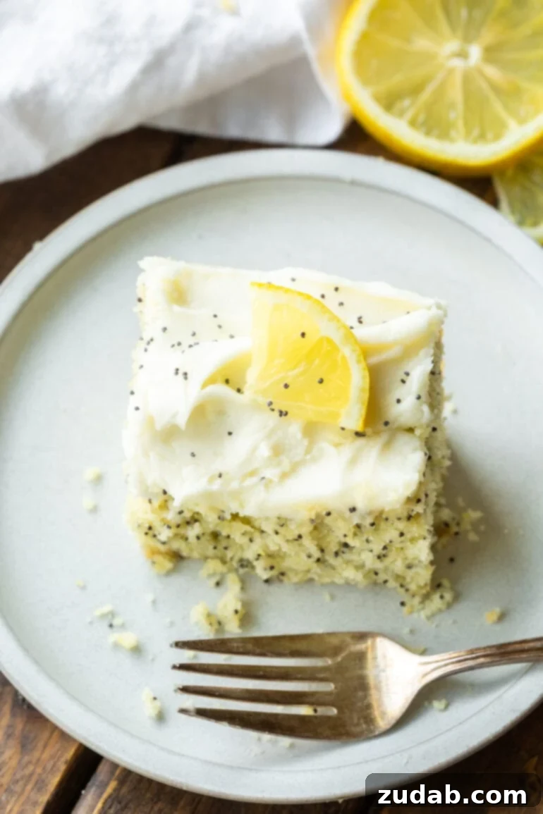 a photo of a slice of lemon poppy seed cake sitting on small white dessert plate topped with a smooth cream cheese frosting and a thin slice of lemon