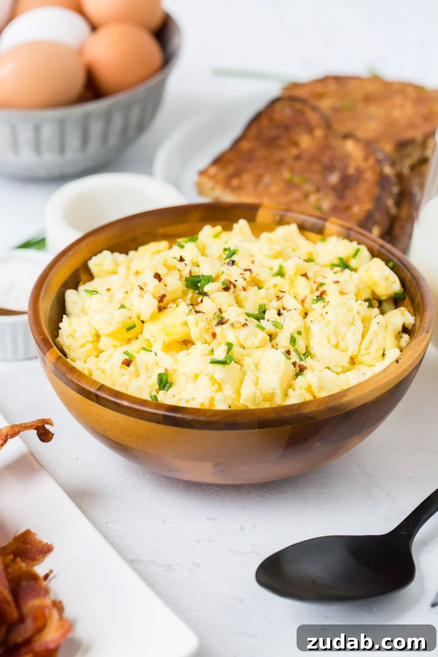 The Best Scrambled Eggs A wooden bowl of scrambled eggs. The eggs are sprinkled with slices of green onion. There are two pieces of toast in the background. There is also a bowl of brown eggs in the background.