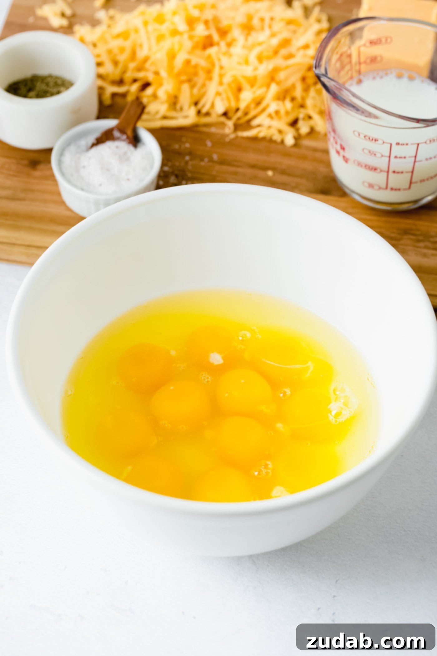 The Best Scrambled Eggs A white bowl with 10 eggs out of the egg shells. In the background is salt, cheese, and milk.
