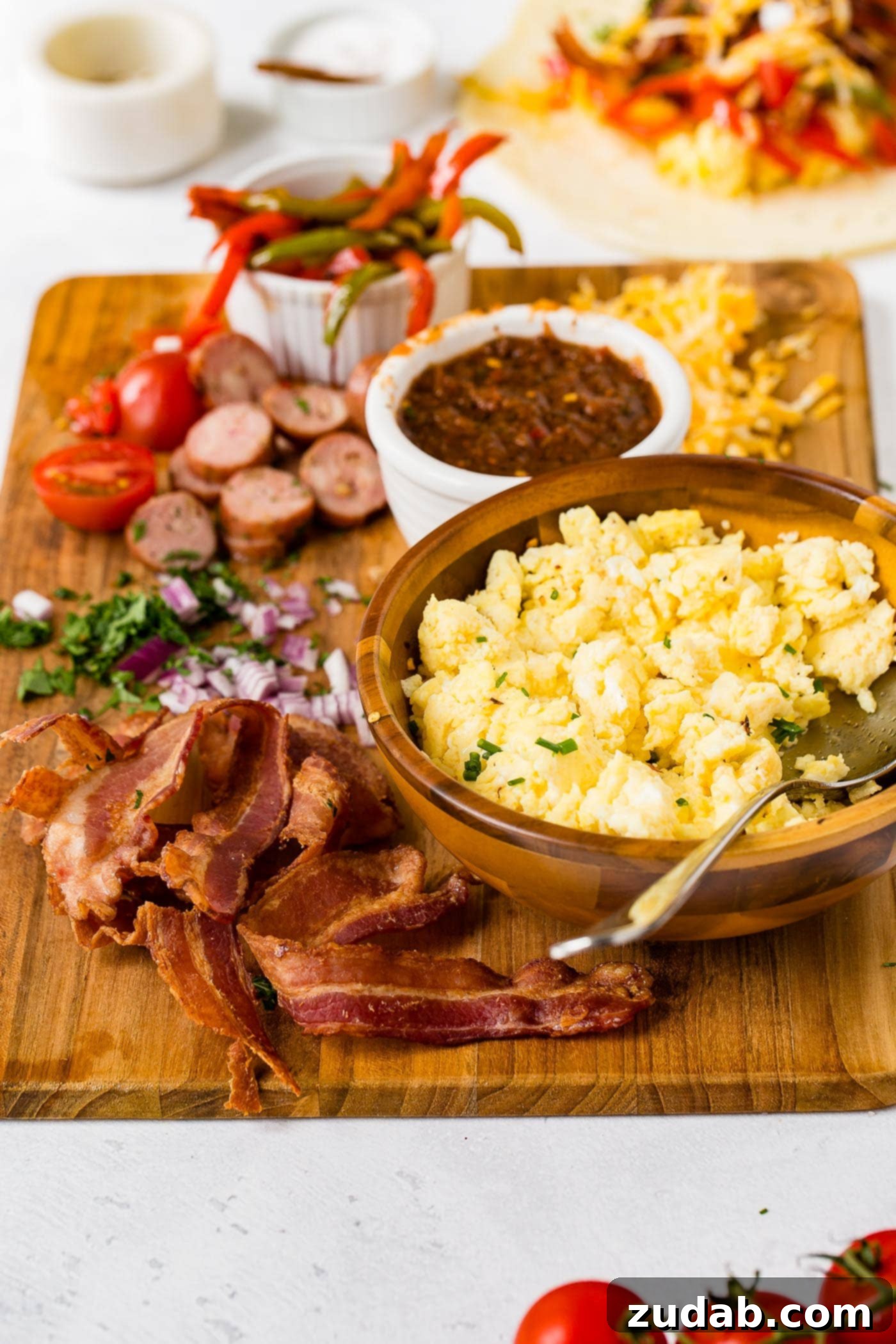 Spiced Apple Sausage Morning Wraps 4 A wooden cutting board holding ingredients for apple chicken sausage burritos. There are sliced sausages, bacon, cheese, peppers, salsa, and a bowl of scrambled eggs with a spoon.