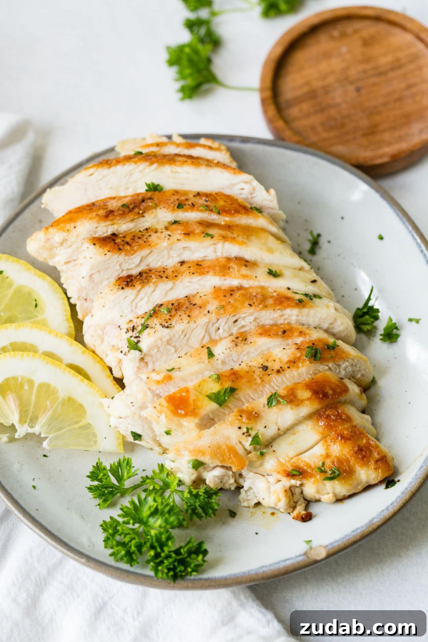 Perfectly Pan-Seared Chicken 3 A white plate with a perfectly cooked chicken breast that has been sliced. There are slices of lemon and a little fresh parsley next to the chicken.