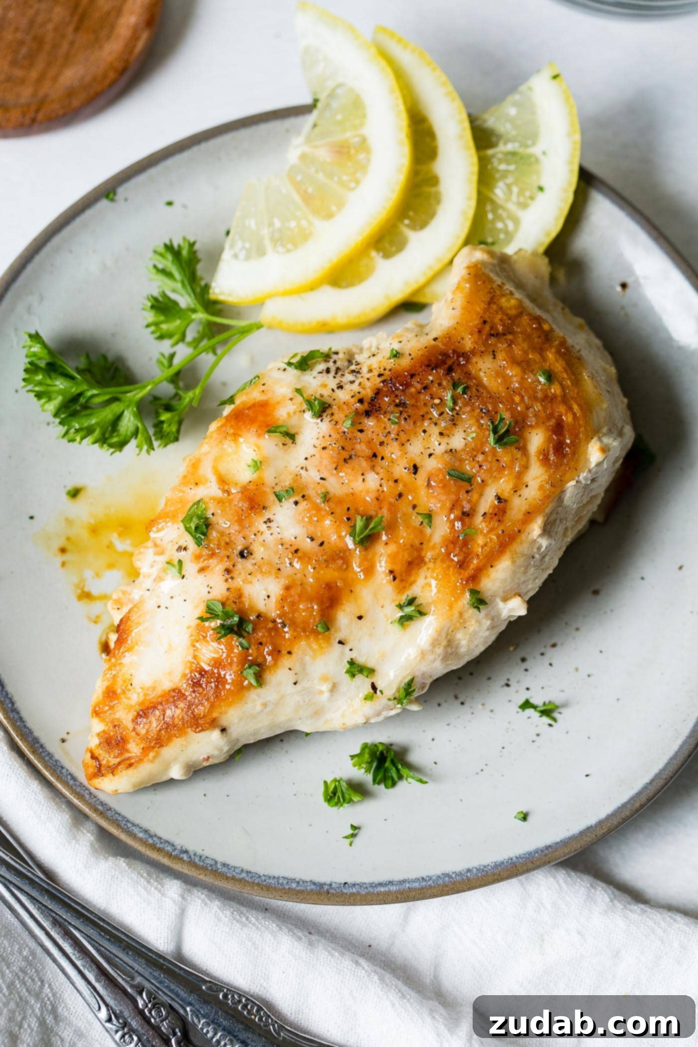 Perfectly Pan-Seared Chicken 4 A chicken breast that has been cooked to a perfect golden brown. The breast is on a white plate with lemon slices and a little fresh parsley.