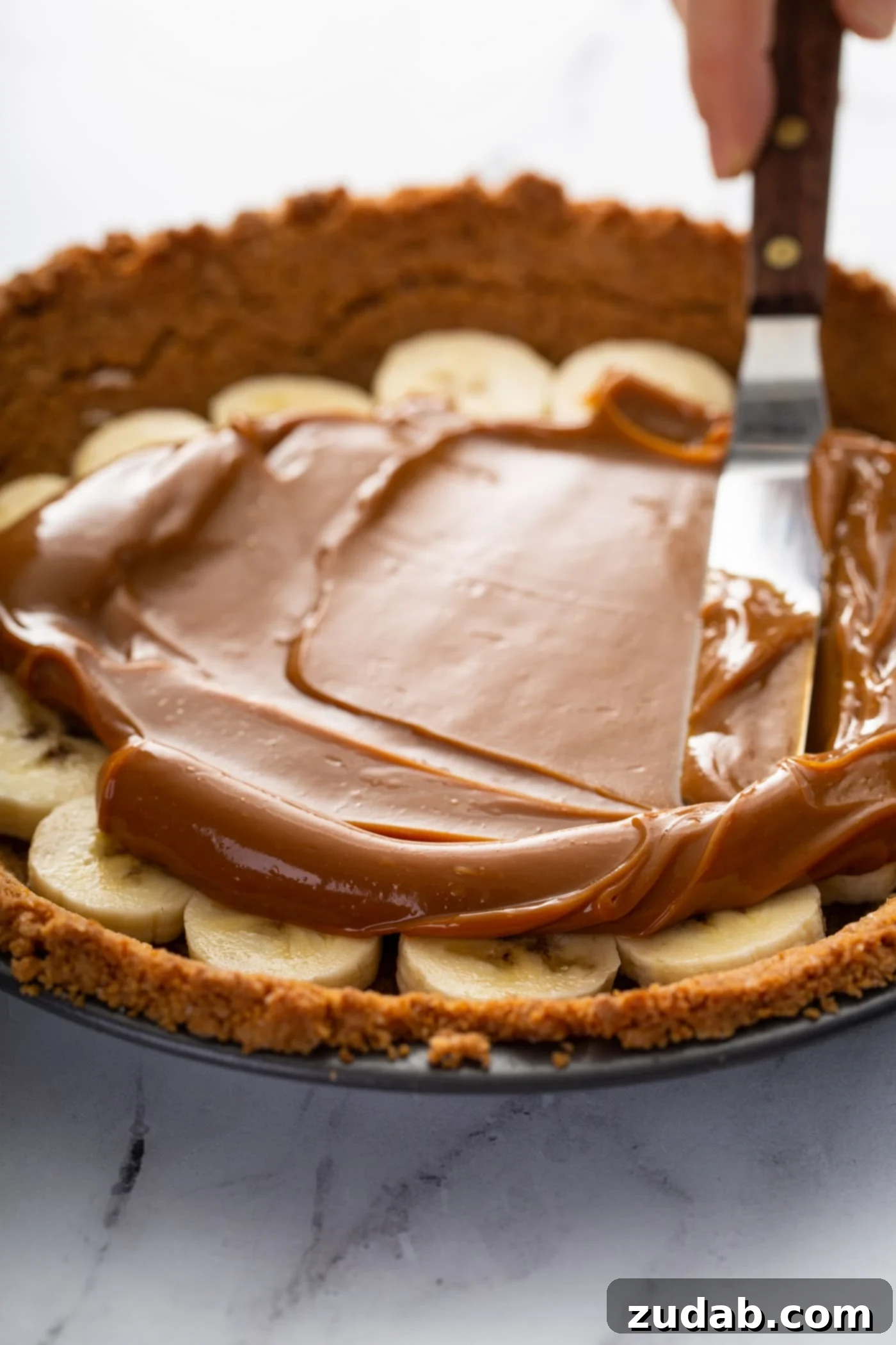 Decadent Dulce De Leche Banana Cream Pie 15 A graham cracker crust in a pie dish. There are sliced bananas in the bottom of the crust and dulce de leche is being spread over the top of the bananas.