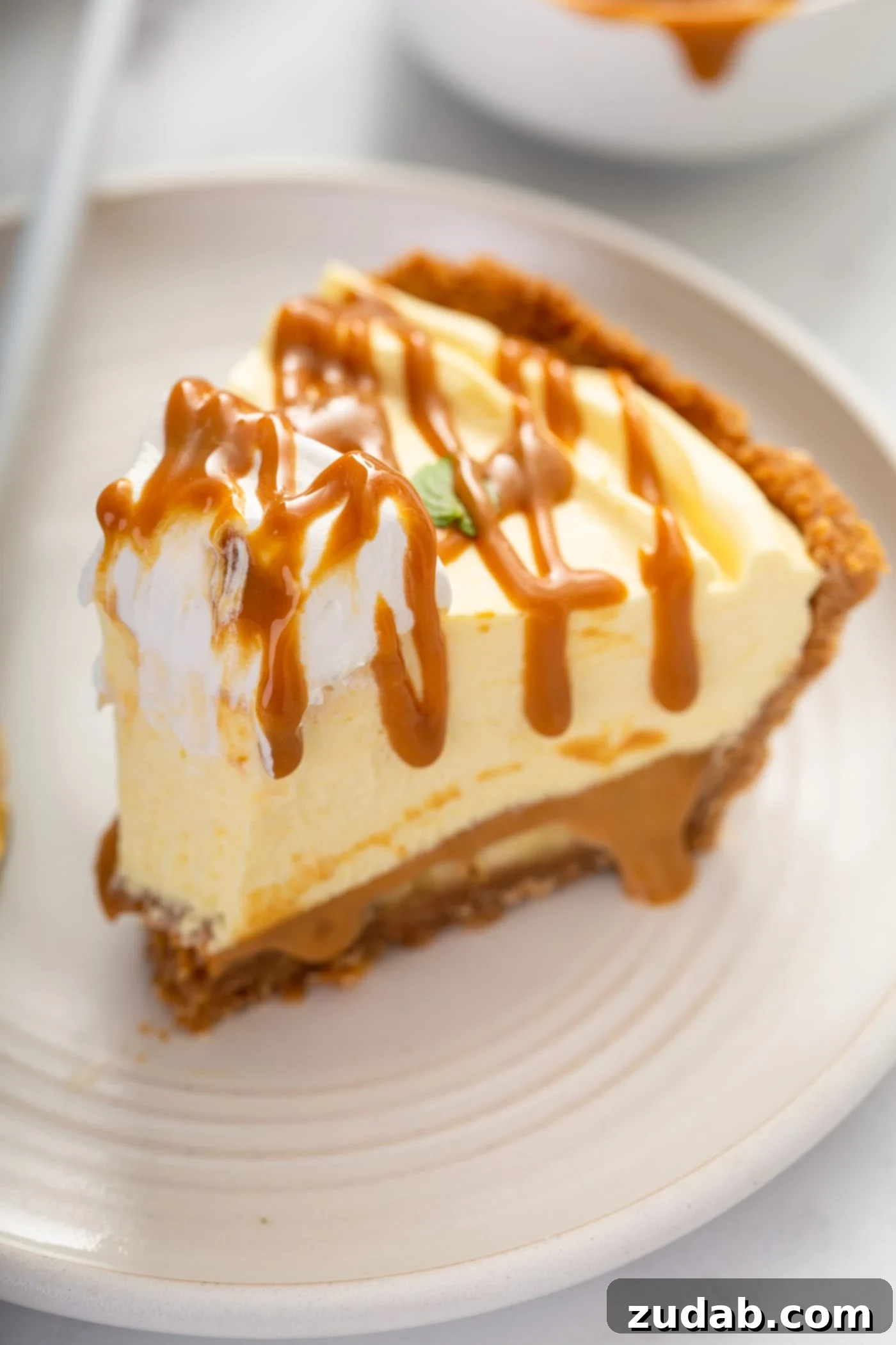 Decadent Dulce De Leche Banana Cream Pie 18 A slice of dulce de leche banana cream pie on a white dessert plate. One bite has been taken from the end of the slice. The pie has a dollop of whipped cream on top and then is drizzled with dulce de leche.