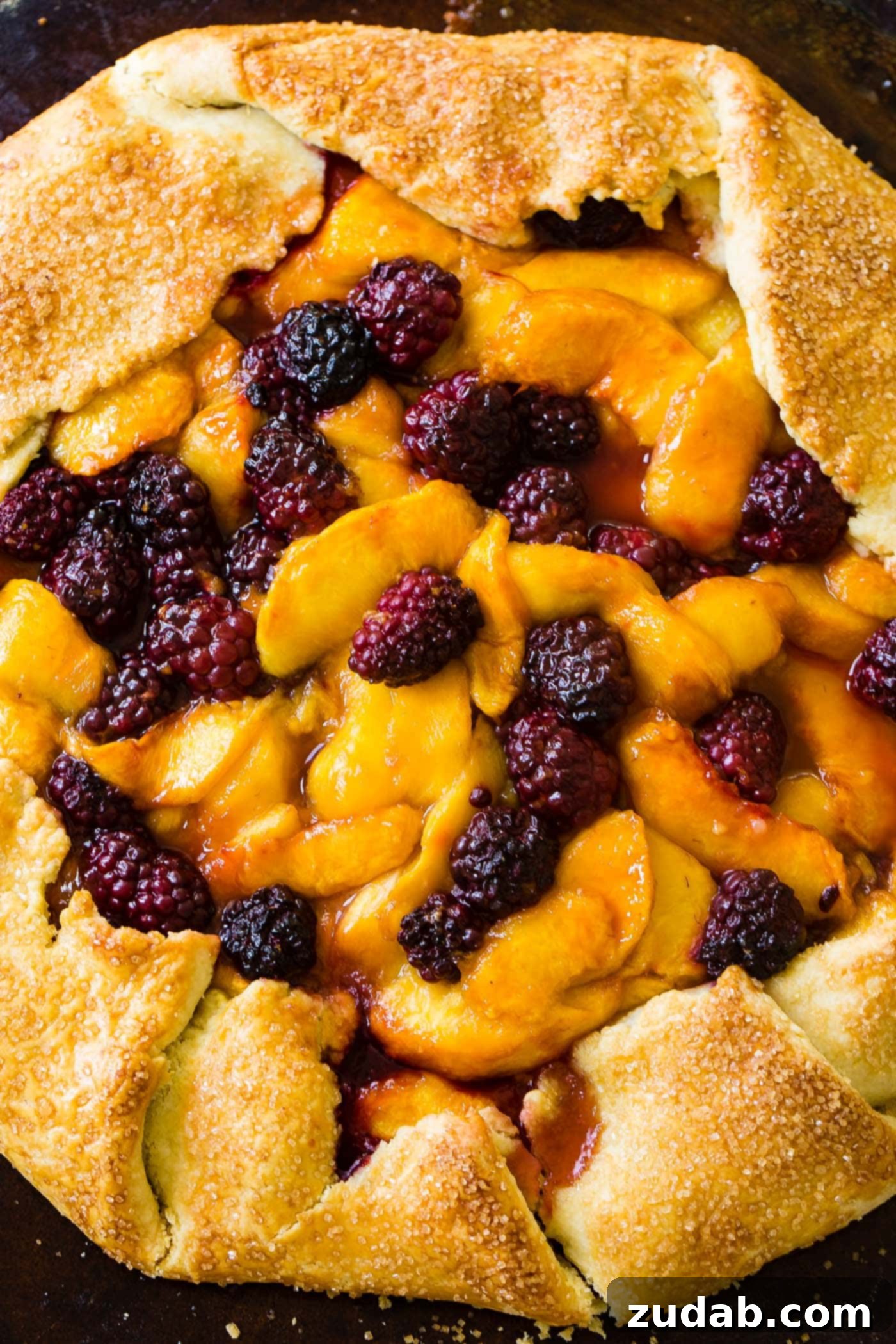 A close-up image of the baked peach blackberry galette, showing the thick, vibrant fruit filling nestled within the golden, rustic crust. The texture of the peaches and blackberries is clearly visible, with some juices bubbling to the surface, indicating a perfectly cooked dessert.