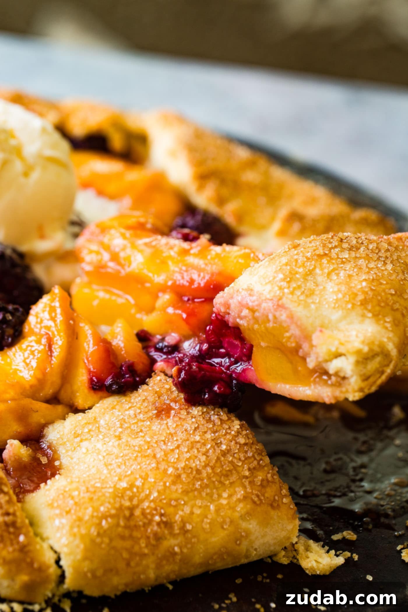 A slice of the rustic peach blackberry galette served on a white plate, revealing the juicy fruit filling and the golden, flaky crust. The background shows the whole galette, reinforcing its homemade appeal. It's a tempting individual serving.