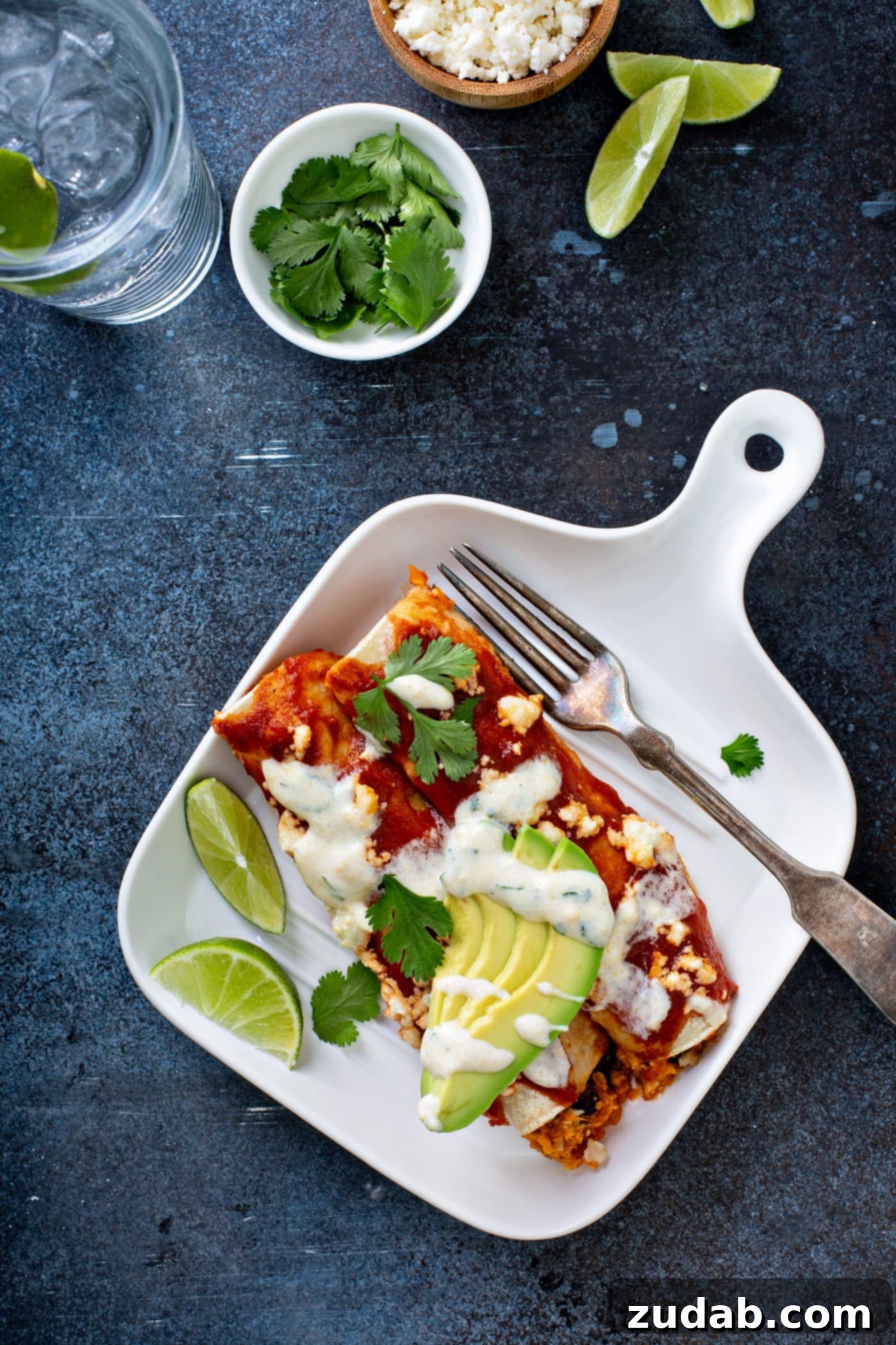 A dinner plate with sweet potato enchiladas and a fork. the enchiladas are topped with red enchilada sauce, sliced avocado, cilantro leaves, and a drizzle of sweet corn crema. There are two lime wedges next to the enchilada. A cup of cilantro and a glass of water are in the background.