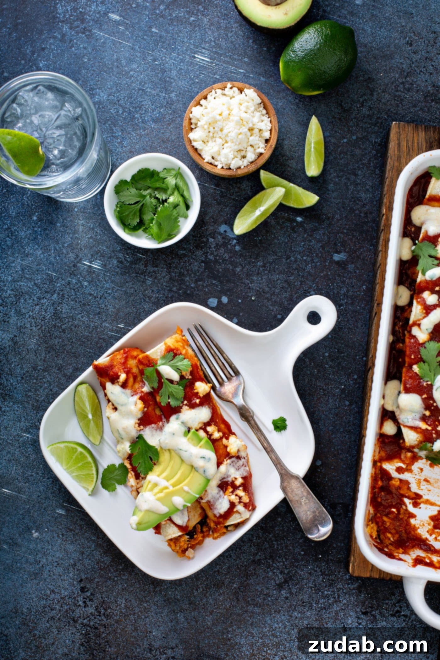 A serving of sweet potato enchiladas on a dinner plate with a fork. The enchiladas are topped with red enchilada sauce, queso fresco cheese, sliced avocados, cilantro, and a drizzle of sweet corn crema. A baking dish of enchiladas and garnishes are in the background.