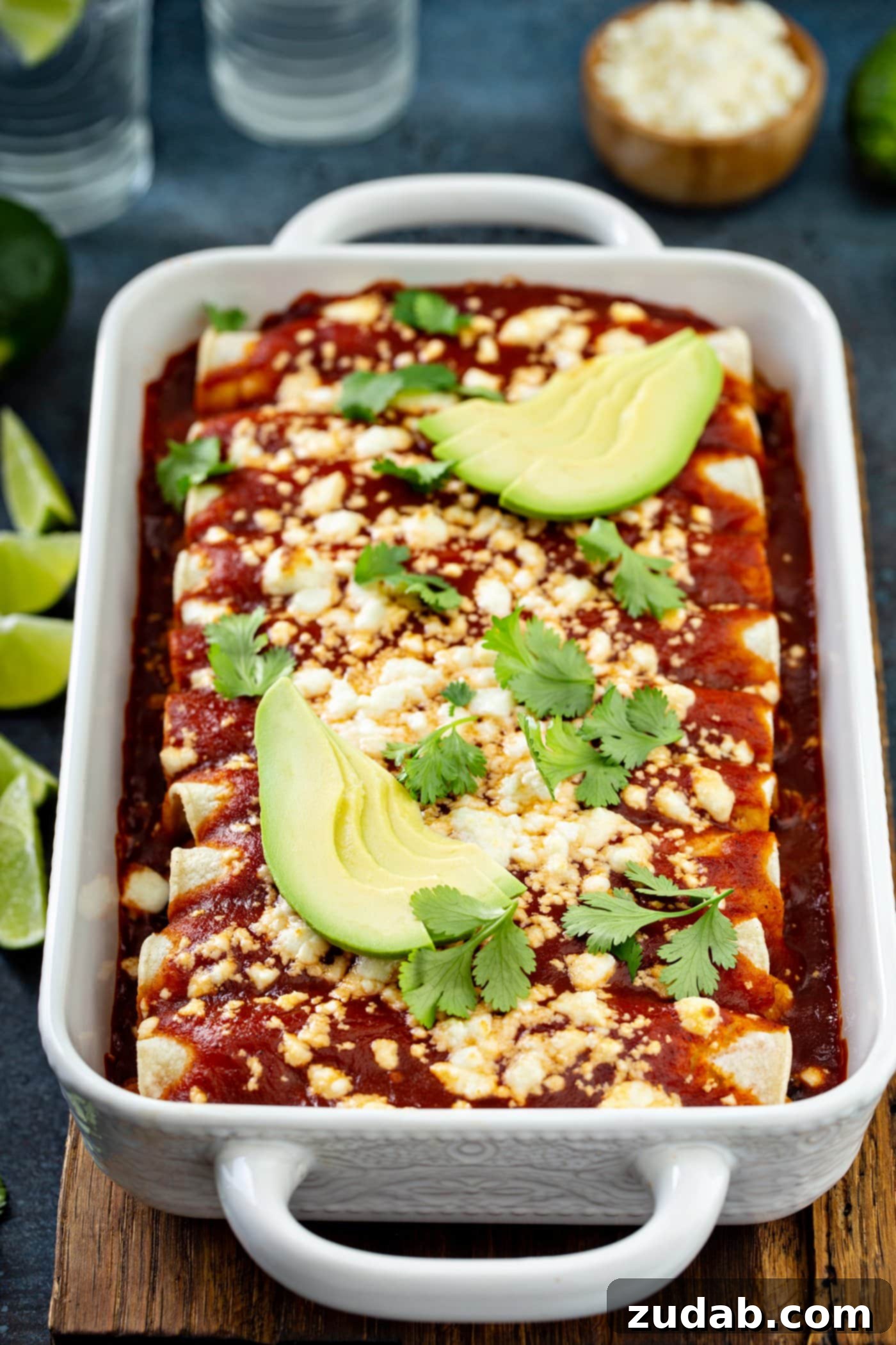 A casserole dish filled with sweet potato enchiladas, topped with red sauce, queso fresco, avocado slices, and cilantro leaves.