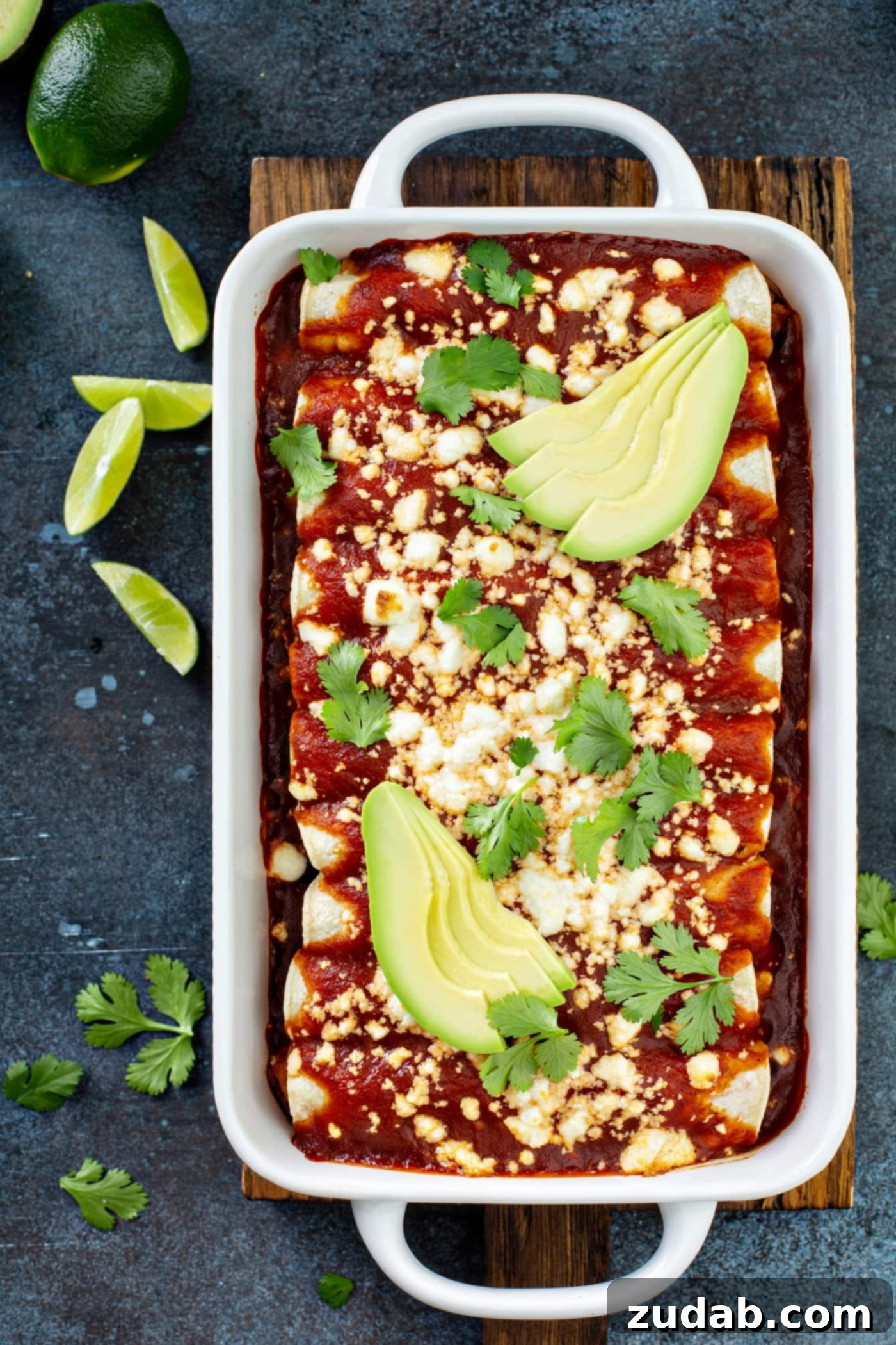A white casserole dish with sweet potato enchiladas, covered in red enchilada sauce and sprinkled with queso fresco cheese, avocado, and cilantro leaves. Lime wedges and cilantro are scattered on the table.