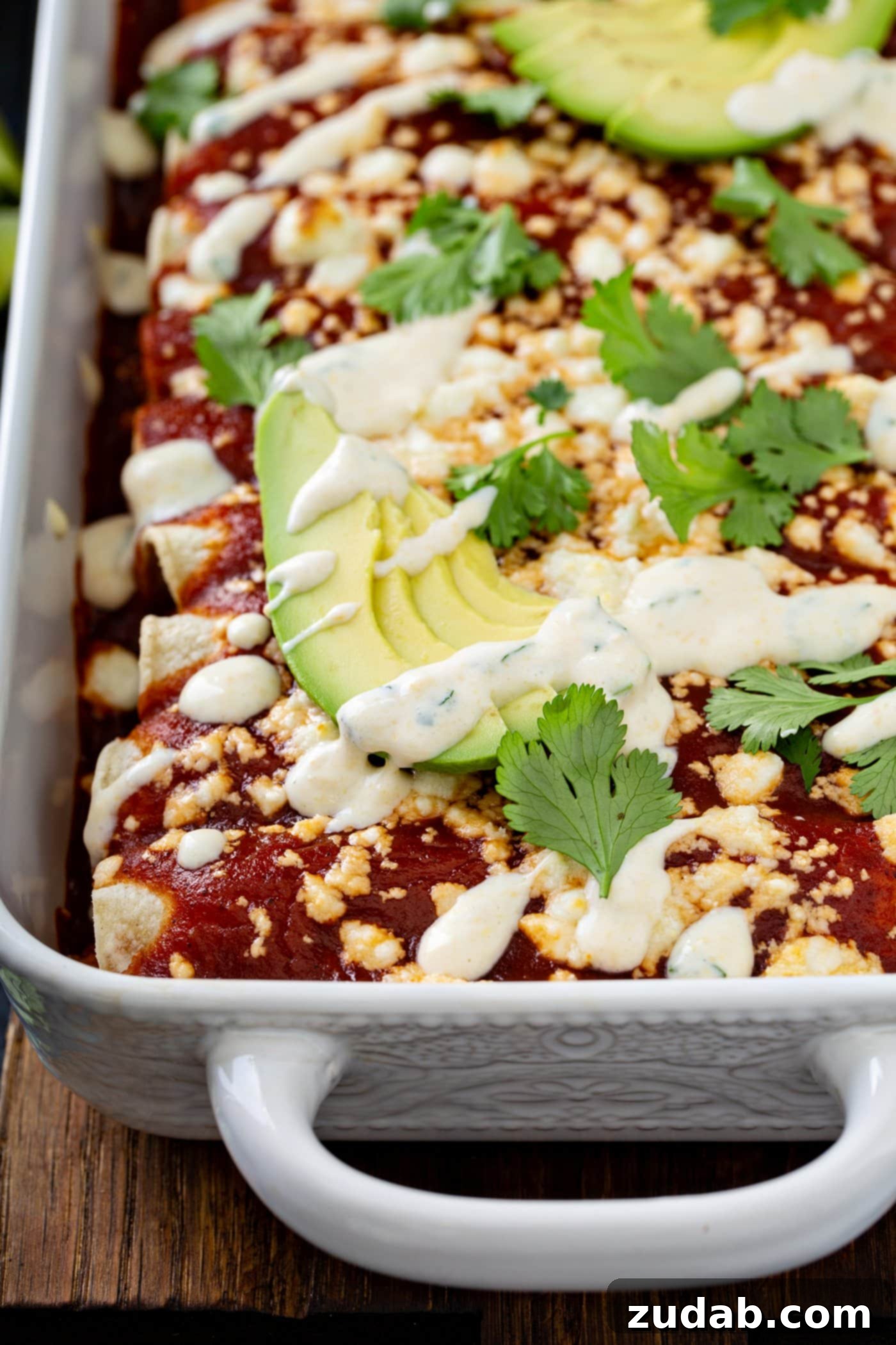 A close-up of a baking dish of sweet potato enchiladas. They are topped with a glossy red enchilada sauce, crumbled queso fresco cheese, fresh avocado slices, and vibrant cilantro leaves, with a delicate drizzle of sweet corn crema finishing the presentation.