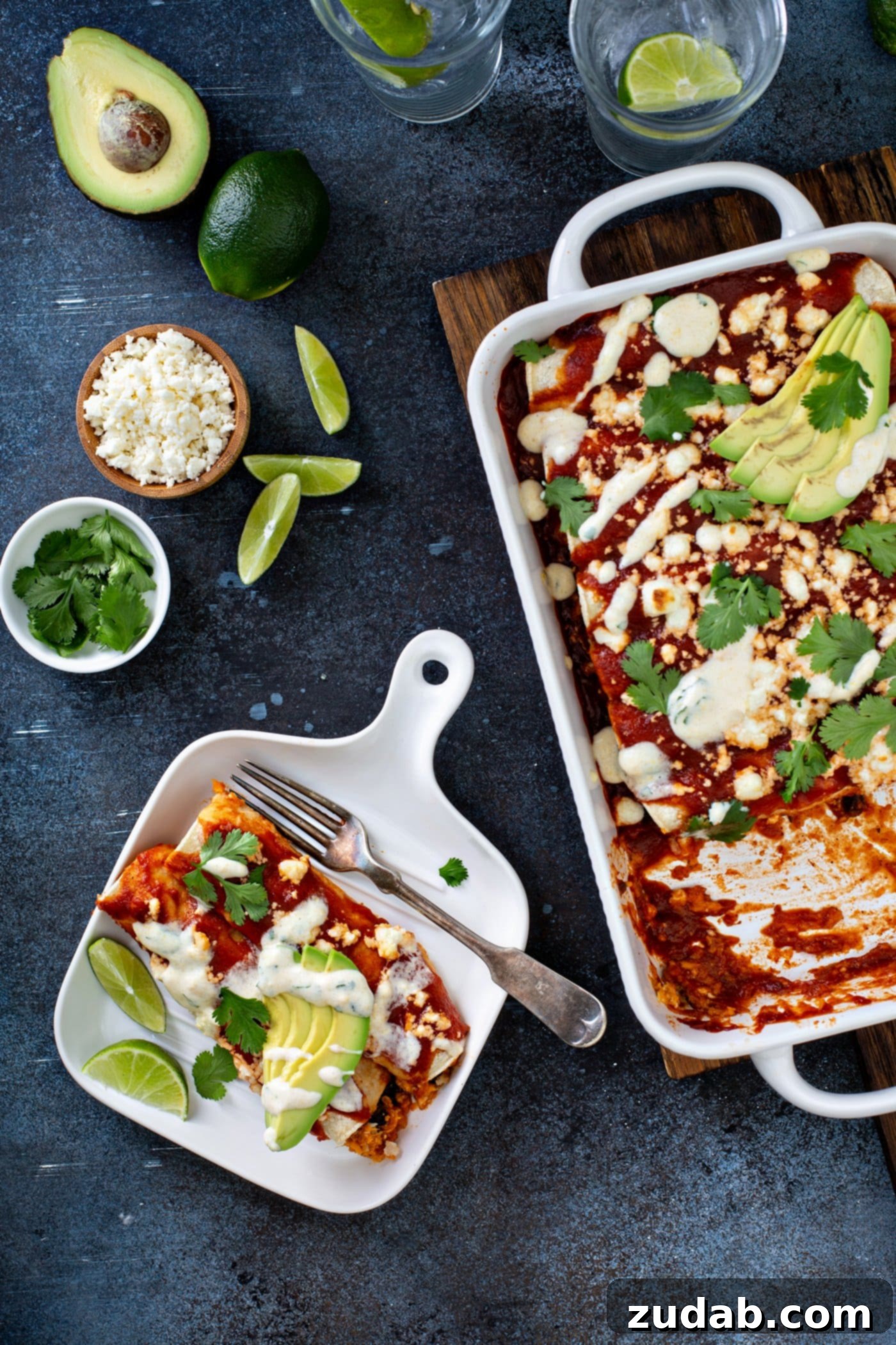 A dinner plate with a serving of sweet potato enchiladas. They feature a red enchilada sauce, sliced avocado, cilantro leaves, and crumbled queso fresco cheese on top, with a delicate drizzle of sweet corn crema. A fork rests on the plate, and a casserole dish with the remaining enchiladas is in the background, alongside lime wedges, a bowl of cheese, and fresh cilantro.