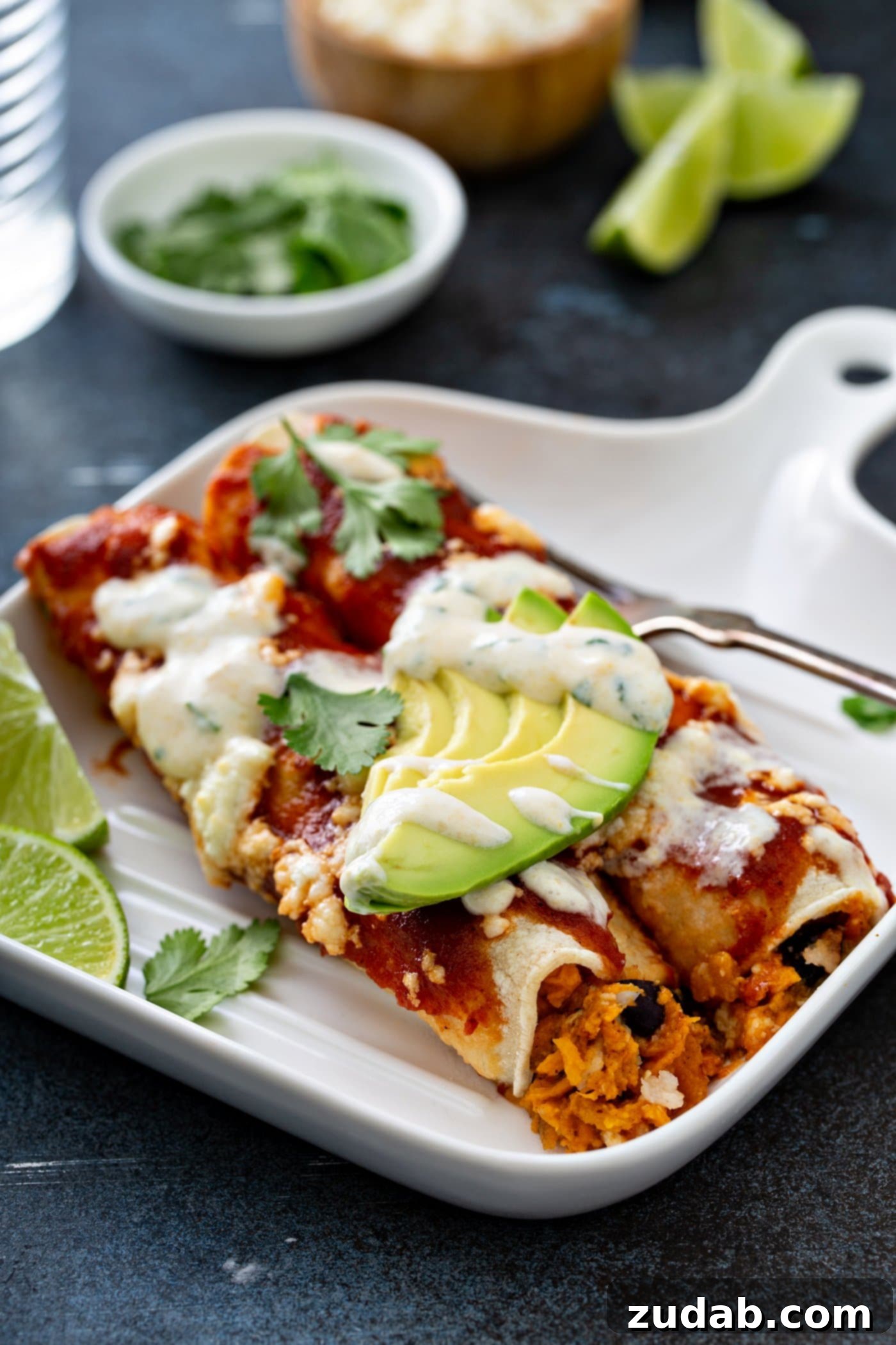 A dinner plate with two sweet potato enchiladas. A fork and two lime wedges are on the plate. The enchiladas are beautifully garnished with red enchilada sauce, crumbled queso fresco cheese, fresh avocado slices, and cilantro leaves, finished with a sweet corn crema drizzle.